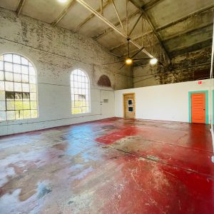Spacious room with high ceiling, large windows, red faded floor, and exposed beams.