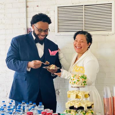 Two people smiling, sharing cake at a table with drinks and cupcakes.