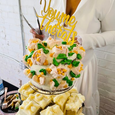 Person in white holds a decorated cake with floral design and gold topper above a platter of frosted cake squares.