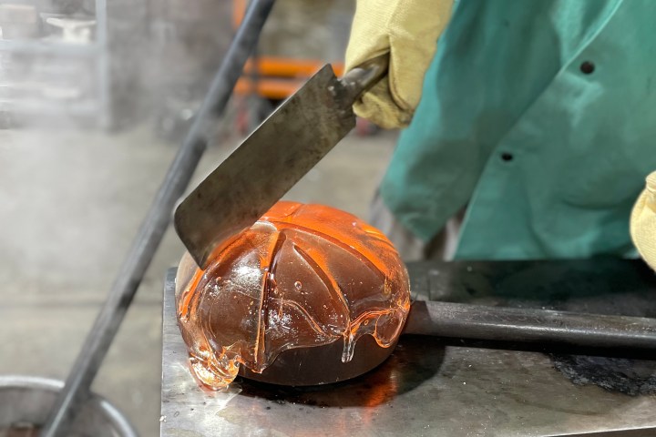 Person cutting hot glass pumpkin on steel table in workshop.